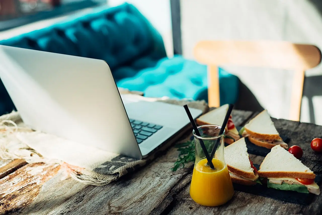 A cozy workspace features a laptop next to a tasty sandwich and refreshing juice on a rustic wooden table. Sunlight streams through a nearby
