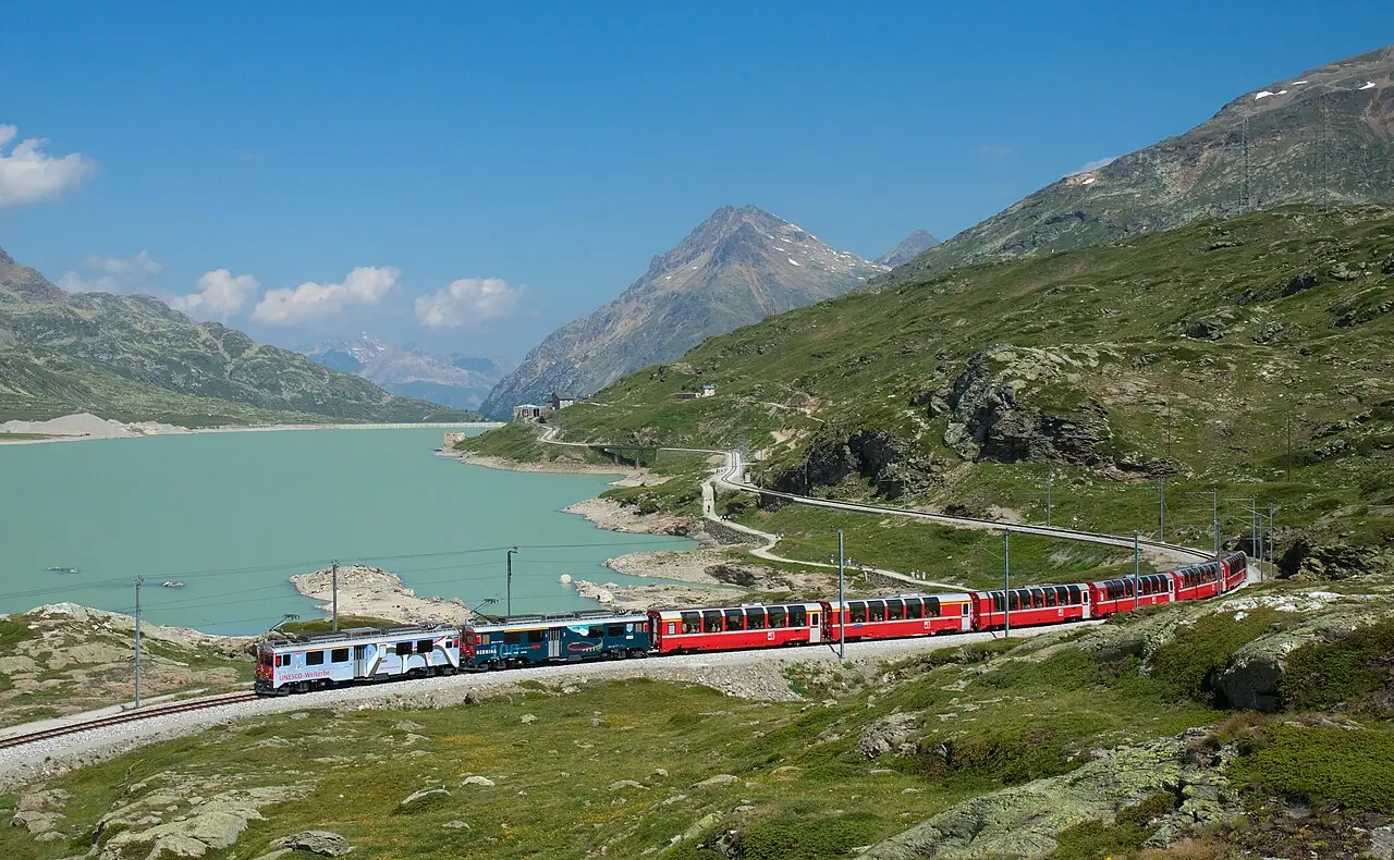 Two RhB ABe 4/4 III motor coaches in multiple unit with a Bernina Express train on the Bernina line , passing Lago Bianco with Ospizio Berni